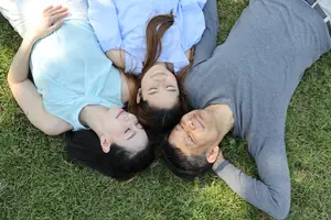 Three people laying on a grass field in a park with their eyes closed and smiling