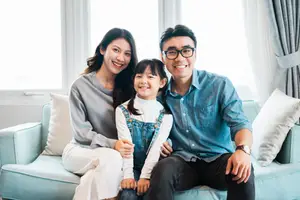 A family of three, a woman, a man, and a little girl, are sitting on a couch with pillows, smiling and posing for a photo in their living room.
