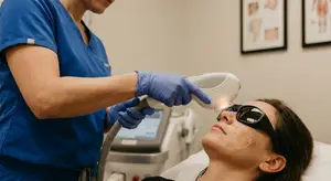 A woman receives laser treatment on her face from a person wearing blue scrubs and gloves.