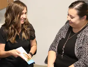 A smiling woman in a black top and leopard print cardigan sits across from another smiling woman, both engaged in conversation and holding a magazine with a woman's image on the cover.