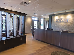 A room with wooden floors, a brown reception desk, a large cabinet with glasses, a chair, a window, and a wall with a sign and a glass door.