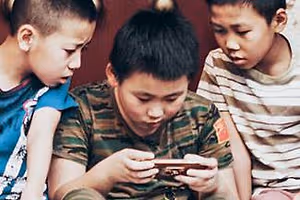 Three young boys are sitting on a couch and looking at their phones. They are all wearing casual clothing, and the boy in the middle has a camouflage shirt with a flag on it. The boy on the left is wearing a blue shirt with a pink design, while the boy on the right is wearing a white shirt with stripes. They are probably looking at a video or a photo on their phones.