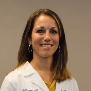 A smiling woman wearing a white lab coat and yellow shirt, standing in front of a light-colored wall.