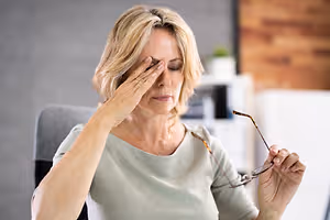 A woman sitting at a desk and rubbing her temples with her eyes closed, holding a pair of glasses in her other hand