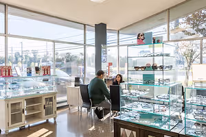 Two people are sitting in chairs in front of a glass cabinet with glasses in an optical shop.
