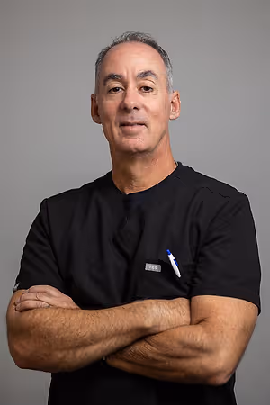 A man wearing a black shirt with a tag on the left side and a pen on the right side, standing in front of a gray background with his arms crossed and smiling at the camera