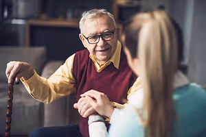 An elderly man holding a cane and sitting on a couch with a woman in front of him