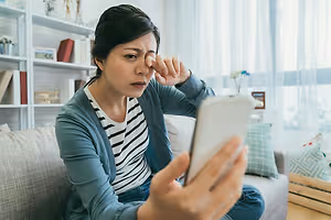 A woman is sitting on the couch, looking at her phone, and rubbing her nose with her hand.