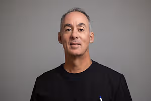 A man wearing a black shirt is smiling and posing for a picture with a gray background.