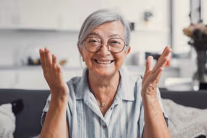 A smiling elderly woman with glasses and a necklace is raising both hands in a room