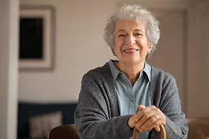 An elderly woman with gray hair smiles and looks forward while standing in a room with a couch and a picture frame on the wall behind her.
