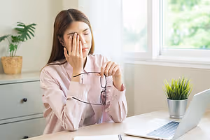 A woman with her eyes closed holding her forehead while sitting in front of a desk