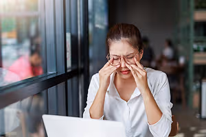 A woman in glasses is holding her forehead with her left hand and looking at something on a laptop in front of her in a coffee shop