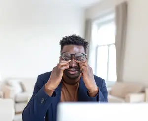 A man wearing glasses and a suit is sitting and holding his face with both hands