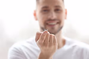 A man wearing a white t-shirt holds a contact lens between his fingers, smiling at the camera.