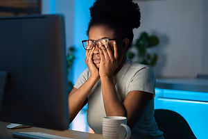A woman is sitting in front of a computer monitor, covering her eyes with her hands, possibly in a dimly lit room.