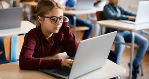 A girl with glasses sitting at a desk using a laptop in a classroom