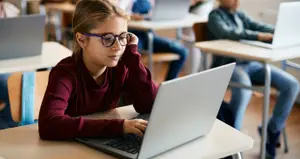 A girl with glasses sitting at a desk using a laptop in a classroom