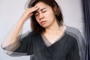 An Asian woman with brown hair is holding her head with her left hand, probably suffering from a headache. She is looking down and seems to be closing her eyes. She is wearing a black blouse. She is standing in front of a white wall, and the light is coming from the right. There is a blue curtain on the right, and it is blurry. 