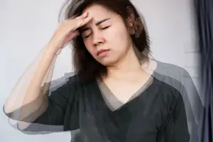 An Asian woman with brown hair is holding her head with her left hand, probably suffering from a headache. She is looking down and seems to be closing her eyes. She is wearing a black blouse. She is standing in front of a white wall, and the light is coming from the right. There is a blue curtain on the right, and it is blurry. 