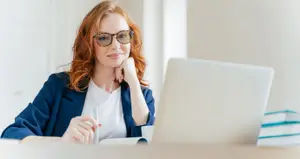 A woman with red hair wearing glasses and a blue blazer is sitting at a desk in front of a laptop, smiling and looking at the camera while holding a pen and a book.