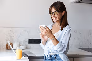 A woman in a kitchen is smiling and looking at her phone.