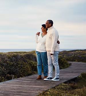 A couple stands on a wooden pathway in a scenic outdoor area near a body of water