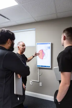 Three men are standing in front of a wall with a monitor displaying an eye image, while the man in the middle is pointing at the monitor with his right hand, possibly explaining something to the other two men. The man on the right is holding a tablet in his right hand, and the man on the left is holding a tablet in his left hand.