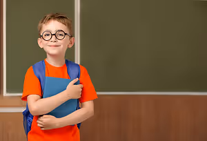 A boy with glasses and a backpack is smiling and holding a book in front of a blackboard.
