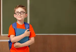 A boy with glasses and a backpack is smiling and holding a book in front of a blackboard.