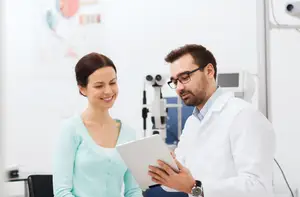 A doctor examines a patient's eyes using an ophthalmoscope while the patient smiles and looks on.