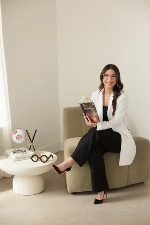 A woman in a white coat and black pants is sitting on a couch and reading a book with the title The Will Eye Manual