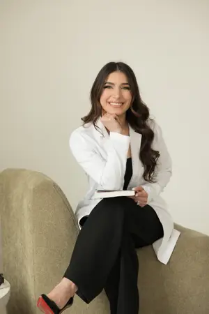 A smiling woman wearing a white coat and black pants is sitting on a couch holding a book.