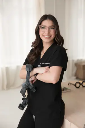 Dr. Shiva Hamade, a female doctor in glasses and a black uniform, is smiling while posing with her arms crossed in front of a desk.