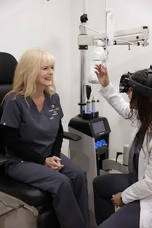 A woman sitting on a chair with a smiling face seems to be talking to another woman, who is standing and holding a virtual reality headset in front of her. Behind them is a white wall, and there is a black machine with some buttons on it.