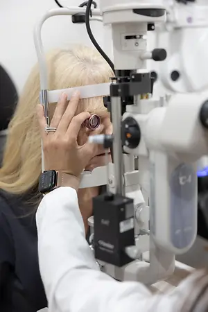 A person with blonde hair is looking into an eye machine with a white light, probably checking her vision at an eye clinic.