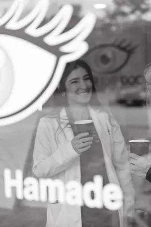 A woman in a white lab coat holding a cup and smiling while standing in front of a glass window with a logo that reads Hamade