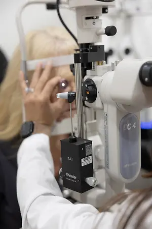 A woman with blonde hair is looking at something while holding her forehead with her right hand and using an ophthalmoscope device with the word 'KAT' written on it. It seems like she is at an optometrist's clinic and is getting her eye examined.