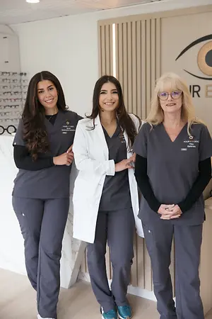 Three female optometry professionals posing in an eye care clinic, with eyeglass displays and an eye logo behind them.