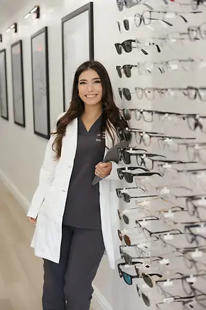 Smiling optometry professional in a white coat standing beside a wall display of eyeglass frames