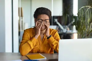 Woman with glasses sitting at desk rubbing her eyes looking stressed