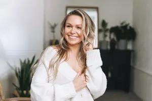 Woman in white sweater standing in a room with plants and a mirror on the wall