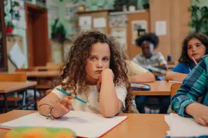 A young girl with curly hair is sitting at a desk in a classroom and seems to be writing something with a pen on a notebook.