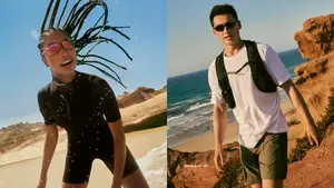 A woman with braided hair and a wetsuit is standing on the beach and a man in a white t-shirt, green shorts, and sunglasses is walking on a cliff next to the sea.