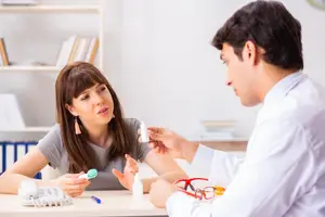 A woman talks to a man wearing a white lab coat in a room with shelves and a desk