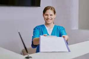 A smiling woman in blue scrubs holding a folder with a paper clip, standing behind a desk with a microphone and monitor in a room