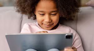 A young girl with curly hair is sitting on a couch and smiling while using a tablet computer.
