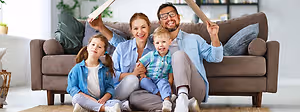 A family of four sits on a couch with two kids on their laps, one holding a book and the other holding a stick