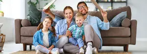 A family of four sits on a couch with two kids on their laps, one holding a book and the other holding a stick