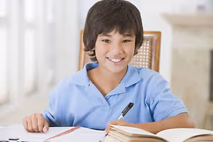 A smiling boy sits at a desk, holding a pen and surrounded by open books and notebooks.